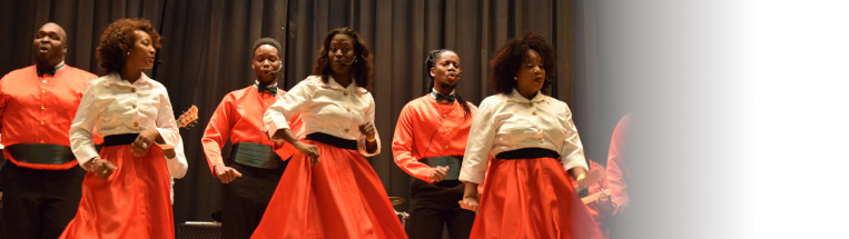 Three Black women in white button ups and orange circle skirts and three black men in orange shirts with black bowties sing in front of a black curtain.