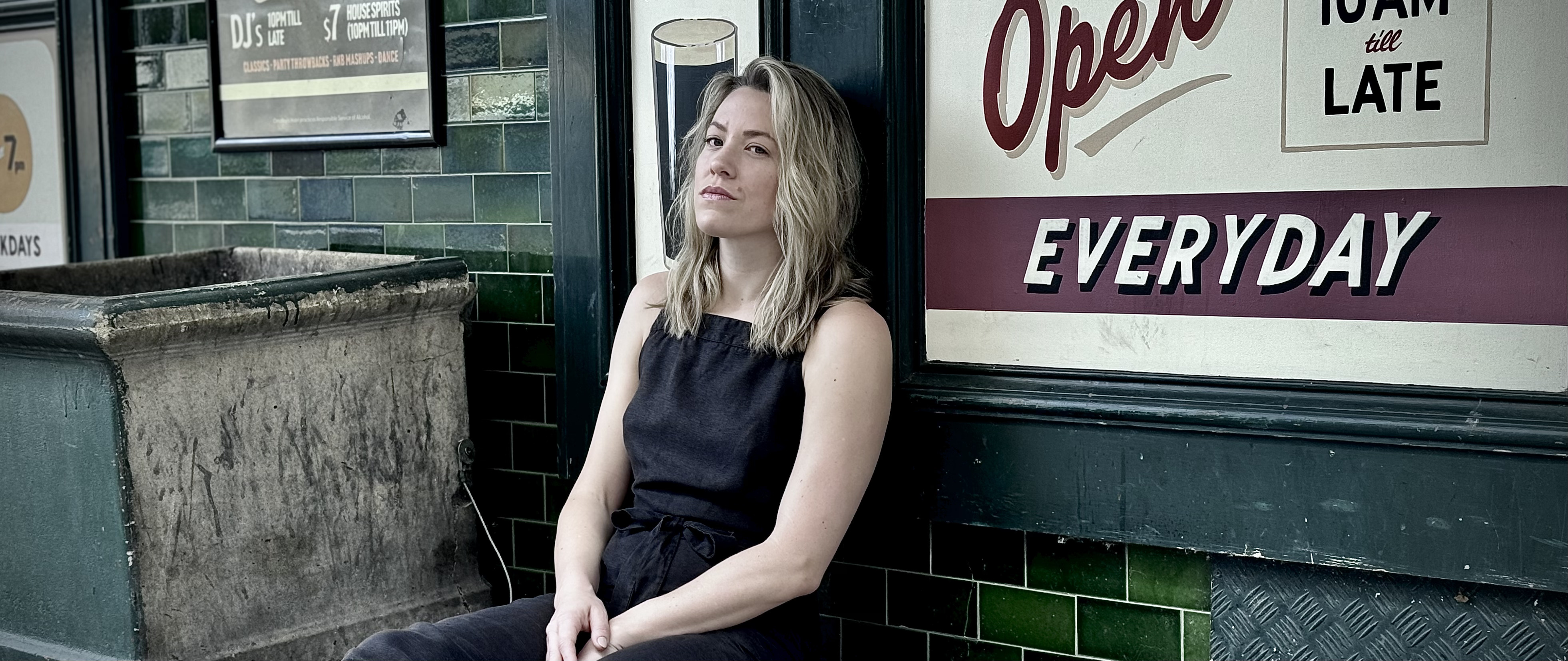 Image for Herself - a white woman with shoulder length blonde hair sits leaning against a wall. She has a picture of a pint of Guiness behind her, and over her shoulder is a sign that says "OPEN" in a stylized font.