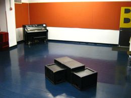 A large room with shiny blue flooring. There is a piano in the corner and a few black rehearsal blocks in the middle of the room.